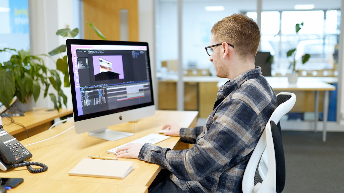 Man sitting at a desk in an office with video editing software open on a computer. 